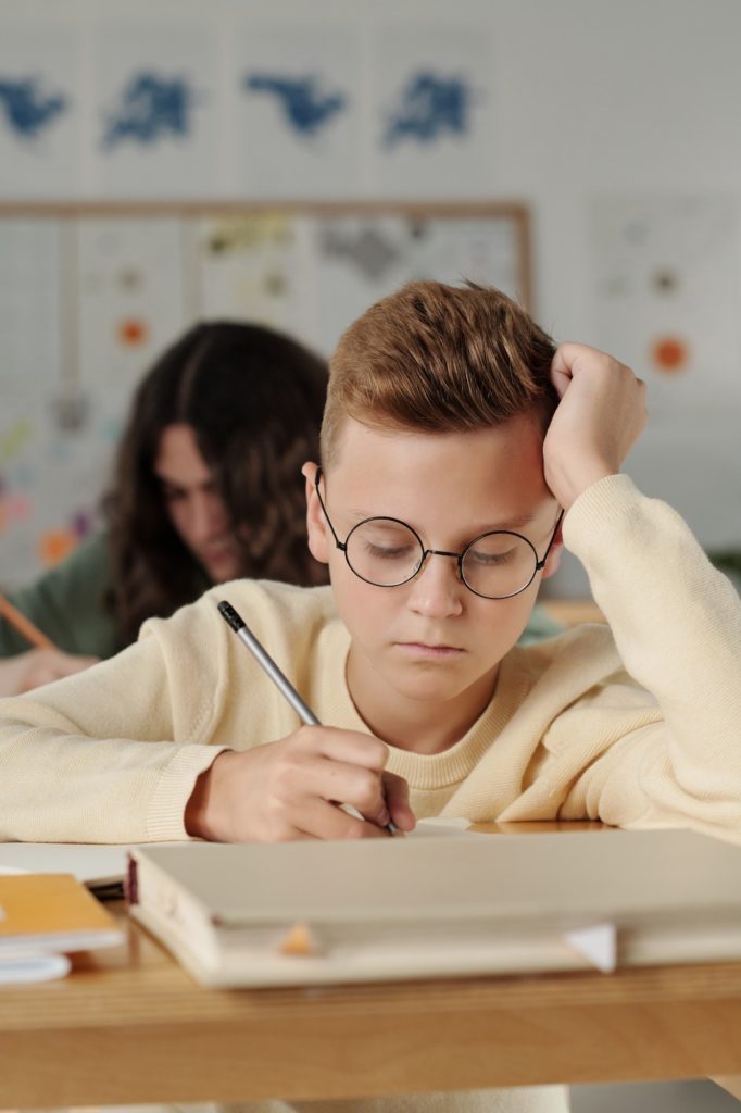 Serious pre-teen schoolboy in eyeglasses making notes in copybook at lesson